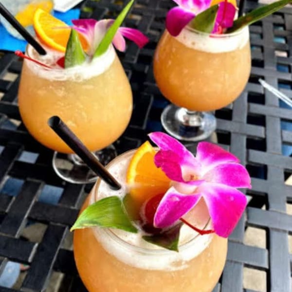 Three tropical cocktails with orange slices, cherries, and pink orchids on a dark patio table.