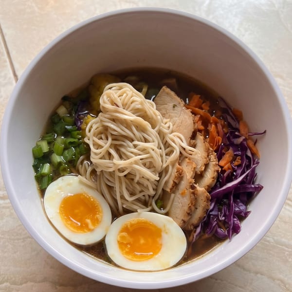 A white bowl of ramen with noodles, sliced chicken, soft-boiled eggs, green onions, and purple cabbage.