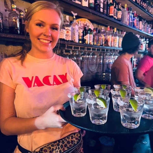 Smiling bartender in a "VACAY" shirt holding a tray of margaritas with lime wedges and salt rims.
