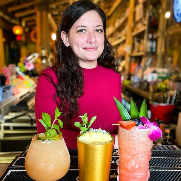 A smiling woman in a red shirt stands behind a bar with three colorful cocktails.