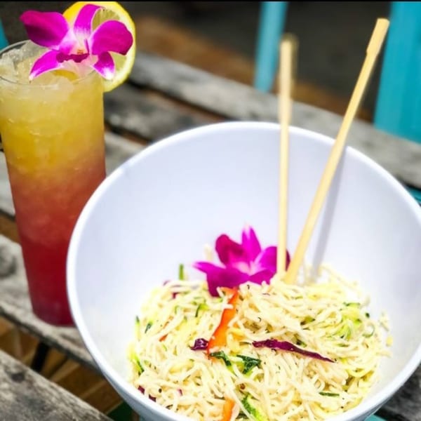 Noodle dish with vegetables and chopsticks next to a tropical drink garnished with an orchid and lemon.