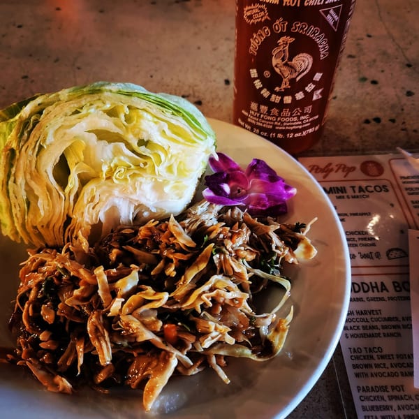 Lettuce wraps with shredded chicken, a purple orchid, and a Sriracha bottle on a table.