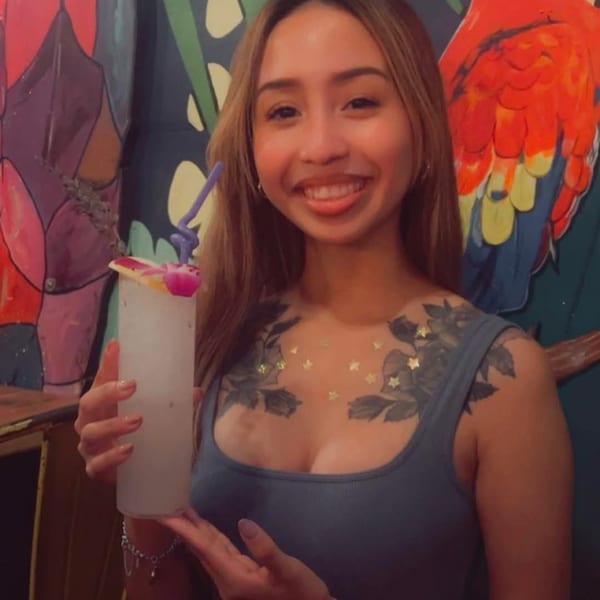 Smiling woman holding a tropical drink with a purple straw, against a colorful mural.