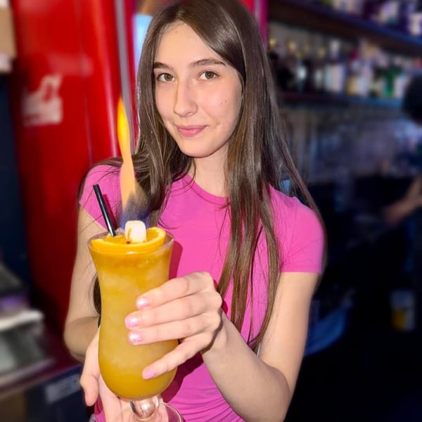 Young woman in a pink shirt holding a flaming orange cocktail with a marshmallow garnish.