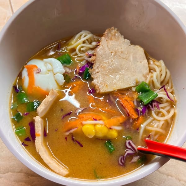 Close-up of a ramen bowl with noodles, chicken, egg, corn, carrots, and red cabbage.