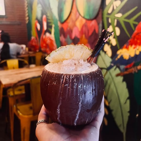 A hand holds a coconut drink with crushed ice, pineapple, and straws, against a vibrant mural.