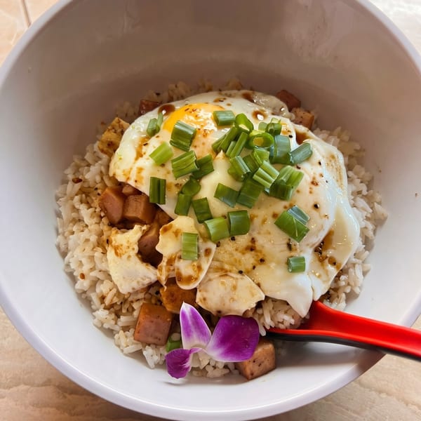 A white bowl of rice topped with fried egg, diced spam, green onions, and a purple orchid.