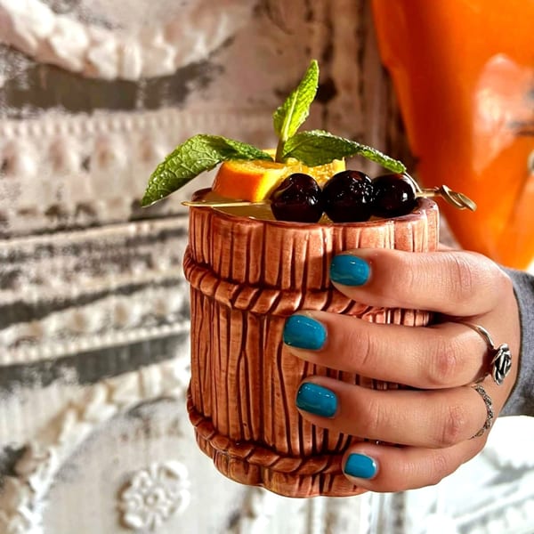 A hand with blue nail polish holds a tiki mug garnished with mint, orange, and cherries.