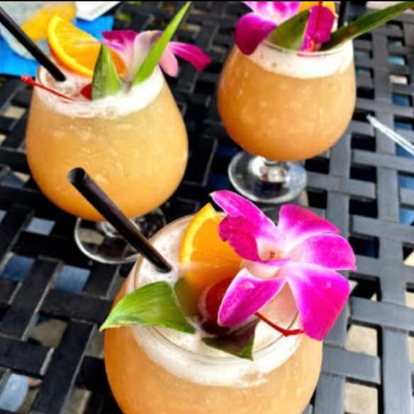 Three tropical cocktails garnished with orchids, orange slices, and cherries on a dark woven table.