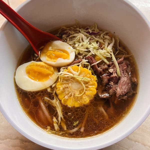 A bowl of ramen with broth, noodles, shredded cabbage, corn on the cob, two soft-boiled eggs, and shredded beef.
