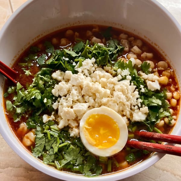 A bowl of pozole with hominy, cilantro, crumbled cheese, and a soft-boiled egg.