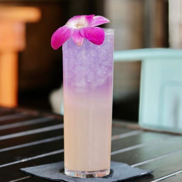 Tall glass with a purple and peach layered drink, garnished with a pink orchid.