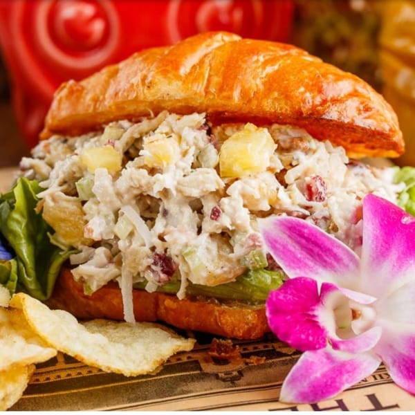 Chicken salad sandwich on a croissant with lettuce, pineapple, and a pink orchid.