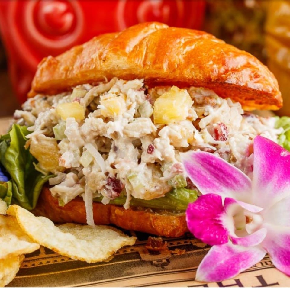 Chicken salad sandwich on a croissant with lettuce, pineapple, and a pink orchid.