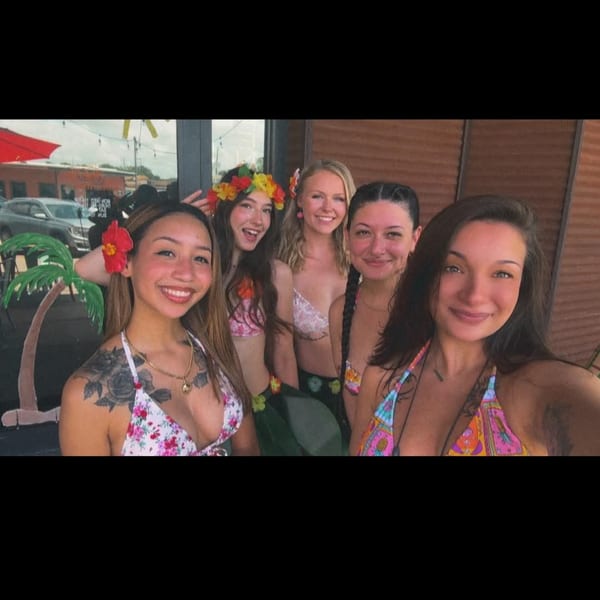 Five smiling women in tropical attire and flower leis pose together for a selfie.