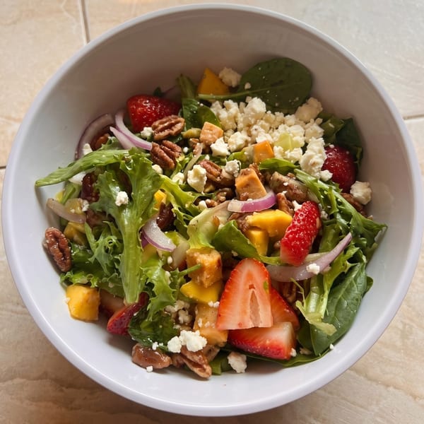 A vibrant salad in a white bowl, featuring mixed greens, strawberries, mango, pecans, red onion, and feta cheese.