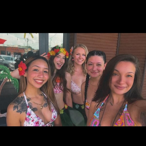 Five smiling women in swimsuits and floral accessories pose together outdoors.