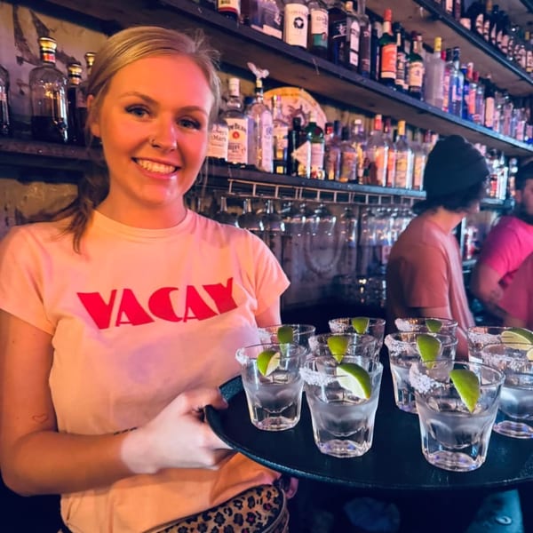 Smiling bartender in a "VACAY" shirt holding a tray of tequila shots with lime wedges and salt.