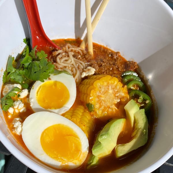 Close-up of a ramen bowl with broth, noodles, ground meat, corn, avocado, jalapeños, cilantro, and two soft-boiled eggs.