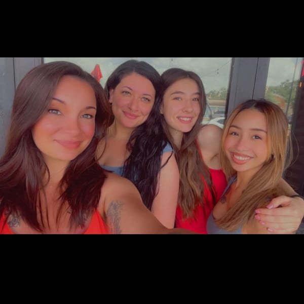 Four smiling young women posing closely together for a selfie outdoors on a sunny day.