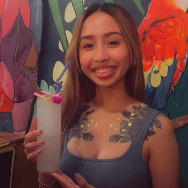Smiling woman holding a tall, frosted cocktail with a purple straw and orchid garnish.