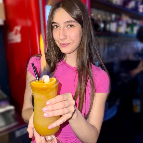 Young woman in a pink shirt holding a flaming orange cocktail with a marshmallow garnish.