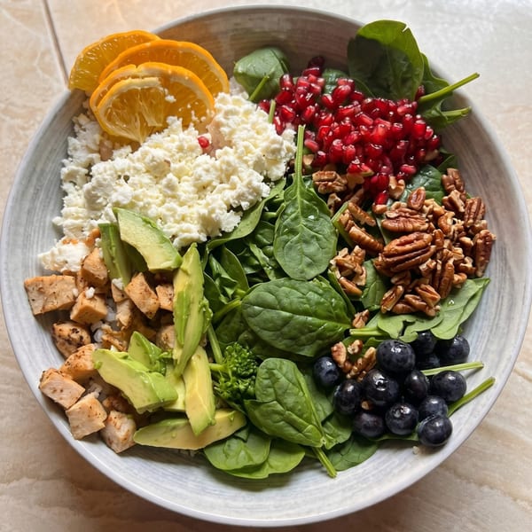 A vibrant salad bowl with spinach, chicken, avocado, feta, oranges, pomegranate, pecans, and blueberries.